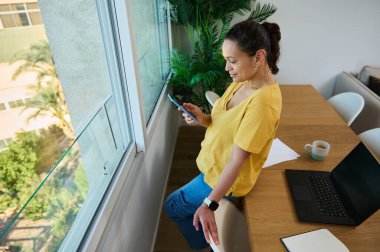 İşten kaçarken akıllı telefon kullanan bir kadın pencerenin kenarına eğilmiş. Ofisinde, yanında bir dizüstü bilgisayar ve kahveyle oturuyor..
