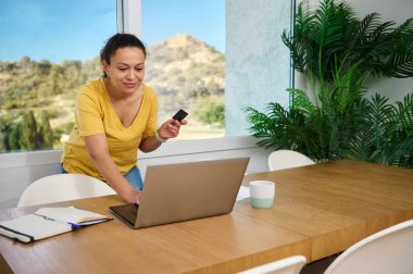 Profesyonel bir kadın bilgisayarını ve kartını rahat ve modern bir ev ortamında aktif online çalışmayı gösteren bir kart kullanıyor. Ev işlerinde odaklanma, iş ve rahatlığı temsil eder.