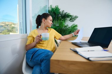 Odaklanmış bir kadın elinde bir kupa, elinde bir dizüstü bilgisayarla tahta bir masada oturan belgeleri analiz eder. Ayarlama, konforlu bir ev ortamı ile mesleki görevler arasındaki dengeyi vurgular.