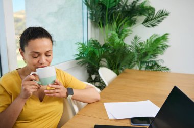 Sıcak bir ev ofisinde, laptopuyla pencerenin önünde çalışırken kahve molası veren neşeli bir kadın. Güneş ışığı ve kapalı alan bitkileri rahatlatıcı ve üretken bir atmosfer yaratırlar..