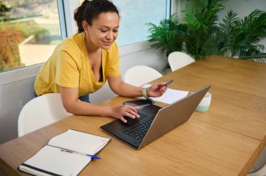 Kendine güveni tam bir kadın. Parlak bir ofiste dizüstü bilgisayar ve kredi kartı kullanıyor. Çalışmalarına odaklanan sahne, modern ticareti ve çevrimiçi alışverişi sıcak, yeşil bir iç mekan ortamında somutlaştırıyor..