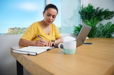 Güneşli bir ofiste çalışmaya odaklanmış serbest çalışan bir profesyonel. Bir dizüstü bilgisayar, defter ve kahve fincanı ile rahat ve verimli bir ortamın tadını çıkarırken görevlerini organize ediyor..