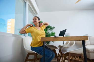 Neşeli bir kadın ev ofisinde uzaktan çalışmaya ara verirken esniyor. Kurulumda, gevşeme ve üretkenlik için ideal bir ahşap masa, dizüstü bilgisayar ve modern dekor yer alıyor..