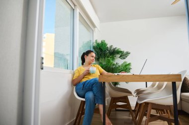 Bir kadın, dizüstü bilgisayarı üzerinde çalışırken, modern bir ev içinde kahve molasından zevk alır. Ayarlama, konforlu ve aydınlık bir çalışma alanında rahatlama ve üretkenlik taşır.