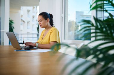 Parlak bir ev ofisinde çalışan bir kadın, huzurlu ve düzenli bir çalışma alanının keyfini çıkarırken özenle dizüstü bilgisayarı üzerinde çalışır, üretkenliği, odaklanmayı ve profesyonel modern yaşam tarzını somutlaştırır..