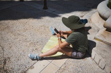 Güneş şapkası takan genç bir gezgin güneşli bir plazada akıllı telefonundan bahsederken haritada geziniyor. Kentsel çevreyi keşfediyor, modern macera ve keşif ruhunu somutlaştırıyor..