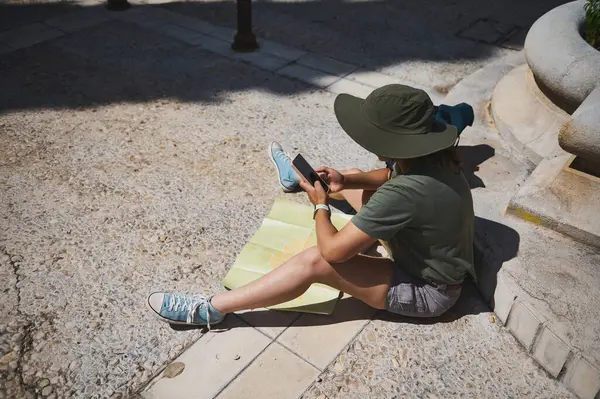 Güneş şapkası takan genç bir gezgin güneşli bir plazada akıllı telefonundan bahsederken haritada geziniyor. Kentsel çevreyi keşfediyor, modern macera ve keşif ruhunu somutlaştırıyor..