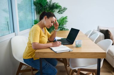 Profesyonel bir kadın dizüstü bilgisayar ve defteriyle masada oturur, işine konsantre olur, özverisini gösterir ve ev ofis ortamında odaklanır..