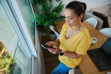 Akıllı telefonuyla meşgul olan kendine güvenen bir kadın ofisinden modern iletişimi ve uzaktan çalışmayı ön plana çıkarıyor. Kişisel dengesi ve teknolojisi olan sakin ve üretken bir ortam..