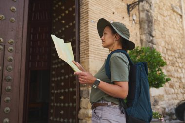 Elinde harita olan bir gezgin, tarihi ahşap bir kapı ve taş bir duvarın yanında duruyor, sakin ve maceraperest bir tavırla ortaçağ kasabasını keşfediyor..
