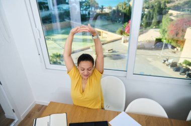 İçerik sahibi bir kadın, evrak ve bilgisayarla dolu bir masada otururken dışarıda sakin bir manzaranın ortasında iş ve rahatlama dengesinin keyfini çıkarıyor..
