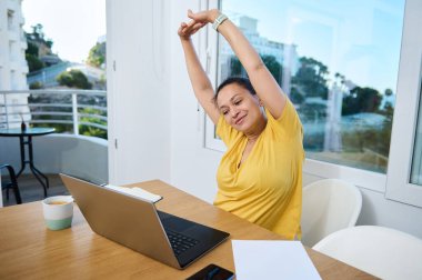 Serbest meslek sahibi bir kadın şık bir ev ofisinde uzaktan çalışma sırasında rahatlar. Canlı sarı giyinerek, kahve molasının tadını çıkarırken esniyor. Dinamik iş dengesine sahip modern yaşam tarzı.