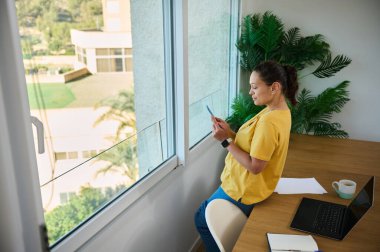 Evde çalışan bir iş adamı, dizüstü bilgisayar ve akıllı telefon kullanıyor. Rahat çalışma alanı doğal ışıklandırma, ev bitkileri ve üretkenliği destekleyen sakin bir atmosfere sahiptir..
