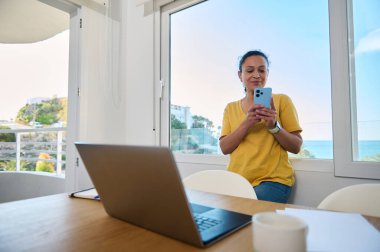 Parlak bir ofiste dizüstü bilgisayar ve akıllı telefonla uzaktan çalışan kendine güvenen bir kadın dijital göçebe yaşam tarzını ve profesyonellik ile açık hava ilhamı arasındaki dengeyi gösteriyor..