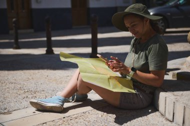 Bir kadın turist elinde haritayla oturmuş, tarihi bir köyü keşfetmek ve güneşin altındaki bir sonraki adımlarını planlamak gibi maceralı bir yolculuğun tadını çıkarıyor..