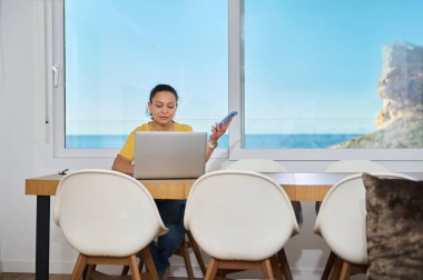 Odaklanmış bir serbest çalışan, dijital sorumlulukları rahatlama anlarıyla dengeleyen, yaratıcılık ve sakinlik arasında modern uzaktan çalışmanın özünü yakalayan manzaralı bir ev ofisinden çalışır..