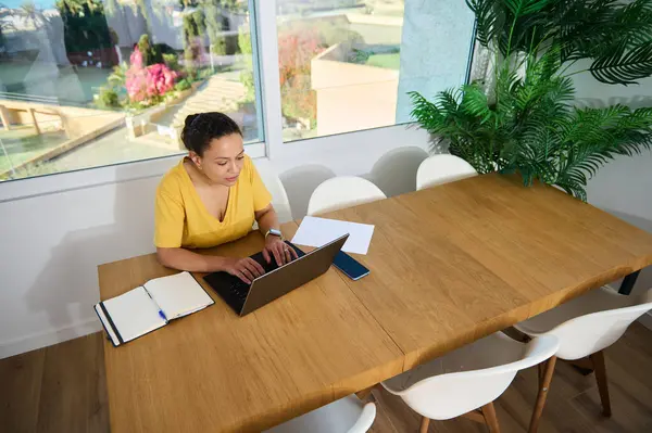 Kendini işine adamış bir kadın parlak, şık bir ev ofisinde dizüstü bilgisayarıyla çalışıyor. Doğal elementler ve modern dekor ile çevrelenmiş, profesyonellik ve rahatlık yayıyor..