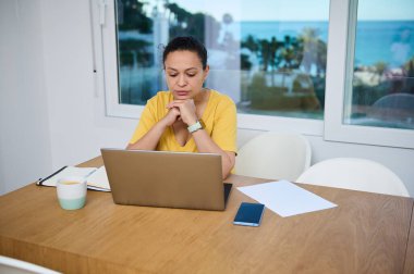 Bir kadın, penceresinden sakin bir sahil manzaralı, profesyonellik ve adanmışlığı aktaran, parlak bir ofis ortamında bilgisayarını uzaktan kontrol ederken dikkatle odaklanır..