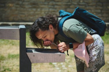 Sırt çantalı bir kadın parktaki bir çeşmeden su içiyor. Sahne sıcacık bir günde susuzluk, ferahlık ve açık hava keşiflerini yakalar..