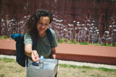 Bir kadın, şehir parkındaki bir çeşmeden su içmek için duraklıyor. Mimari su özellikleri ve yeşilliklerle çevriliyken güneşli günün tadını çıkarıyor..
