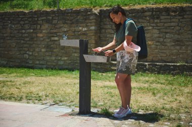 Güneşli bir havada, açık hava çeşmesinde şişesini dolduran bir kadın yeşil bir gömlek ve desenli bir etek giyiyor, taş bir duvar ve çimenlerin yanında..