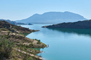 Güzel göl manzarasında huzurlu sular, engebeli kıyı şeritleri ve berrak mavi gökyüzünün altında uzak bir dağ bulunur. Sükuneti ve doğal güzelliği yakalar..