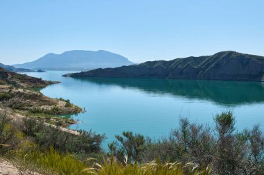 Sakin bir göl, mavi gökyüzünün altındaki tepeleri yansıtıyor. Ön plandaki yemyeşil bitki örtüsü huzurlu atmosferi güçlendirir..