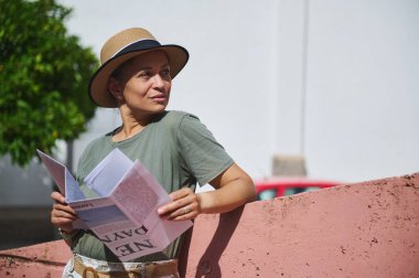 Gündelik giysiler ve hasır şapkalı bir kadın açık havada pembe bir duvarın önünde dururken elinde bir harita tutuyor..