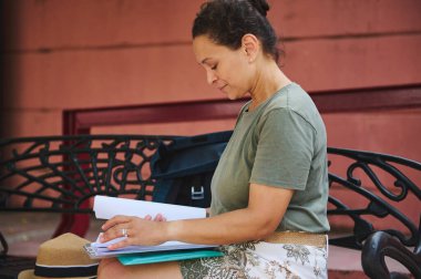 Bir kadın dekoratif bir parkta oturmuş gazeteleri okuyor. Sahne odak ve sükuneti yansıtıyor, yakınlardaki eşyalarla açık hava eğlencesi vurgulanıyor.