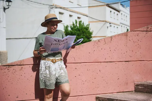 Açık havada, şapkalı bir kadın ve yaz kıyafeti giymiş bir kadın güneş ışığı altında parlak bir duvara gazete okur. Rahatlık ve rahatlık yayar..