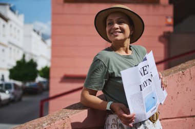 Keyifli bir kadın beton korkuluklara yaslanıyor, yaz şapkası takıyor, elinde gazete tutuyor ve güneşli bir şehir gününün tadını çıkarıyor..