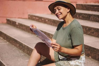 Neşeli bir kadın, güneşli bir günün tadını çıkarır. Merdivenlerde oturur, hafif bir yaz kıyafeti giyer ve broşür okur. Boş zamanları ve mutluluğu vurgulayan parlak bir ortam.