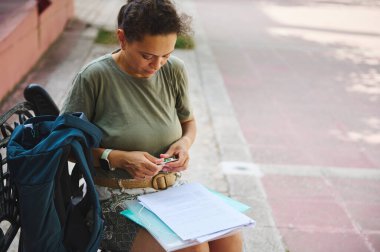 Doğal ışıkla çevrili bir bankta belgeleri düzenlemeye dalmış bir kadın. Odaklanmış ifadesi, günlük çalışma veya akademik faaliyetlerde hazırlık ihtiyacını ve kararlılığını iletiyor..