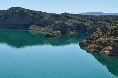 Canlı yeşilliklerle çevrili engebeli kaya oluşumlarına komşu sakin turkuaz bir gölü tasvir eden huzurlu bir manzara, ahenkli bir doğal ortam yaratıyor..