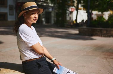 Güneşli bir parkta açık havada oturan, beyaz gömlekli ve şapkalı bir kadının huzurlu görüntüsü. Ayar doğal ışığı yansıtıyor ve huzurlu ve huzurlu bir ortam sağlıyor..