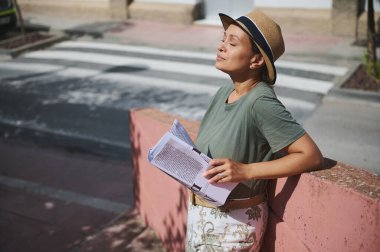 Şık bir kadın şehir kaldırımında güneşli bir günün tadını çıkarır. Şapka takar, elinde kağıtlarla kırmızı bir duvara yaslanır..
