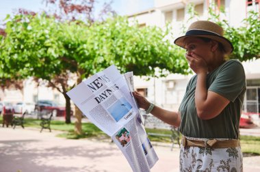Şaşırmış bir kadın, ağaçlar ve güneş ışığıyla dolu bir parkta gazete okurken tepki gösteriyor. Şık bir şapka ve günlük kıyafetler giyiyor..