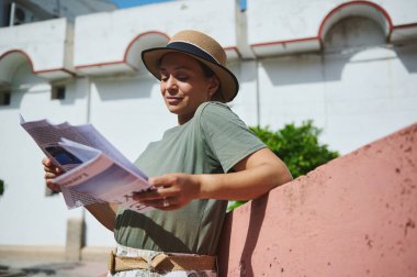 Şapka takan ve günlük kıyafet giyen genç bir kadın güneşli bir günde açık havada dergi okuyor, sıcak havanın ve sakin atmosferin tadını çıkarıyor..