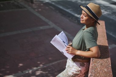 Günlük yeşil gömlekli ve hasır şapkalı bir kadın güneşli bir günde duvara yaslanırken dergi okumaktan zevk alır..