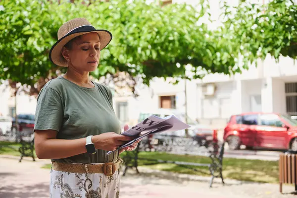 Şapka takan ve günlük kıyafetler giyen bir kadın, ağaçlar ve binalarla çevrili bir şehir parkında dergi okuyor..