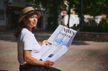 Güneşli bir günde, açık havada parkta rahat rahat oturup, temiz hava ve sükunetin tadını çıkaran şapka takan bir kadın gazete okur..