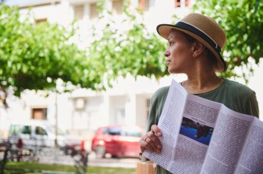 Elinde dergi olan bir şapka takan bir kadın, ağaçların altında sakin bir anın tadını çıkarıyor, doğal ışık ve kentsel elementlerle çevrili..