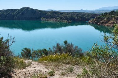 Parlak mavi bir gökyüzünün altında yeşil tepelerle çevrili dingin göl kıyılarda canlı yeşillikler ve sükuneti ve doğal güzelliği temsil eden berrak, yansıtıcı suyla dolu..