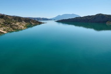 Kurak tepelerle çevrili ve bulutsuz bir gökyüzünün altındaki uzak bir dağ tarafından gözden kaçırılmış, huzur verici mavi sularla dolu bir göl manzarası..