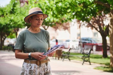 Bir kadın açık havada güneşli bir günün tadını çıkarırken şapka takar, etrafı ağaçlar ve park banklarıyla çevriliyken belge okur, doğayı kentsel sükunetle harmanlar..