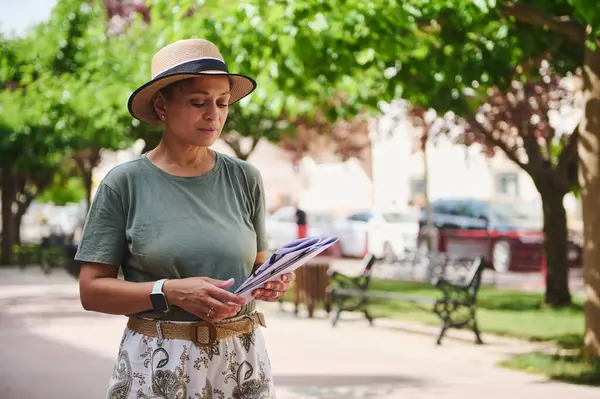Bir kadın açık havada güneşli bir günün tadını çıkarırken şapka takar, etrafı ağaçlar ve park banklarıyla çevriliyken belge okur, doğayı kentsel sükunetle harmanlar..