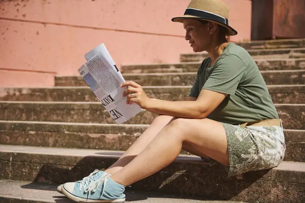 Sıradan giyinmiş bir kadın, dışarıda taş basamaklarda otururken gazete okumaktan, rahatlamaktan ve sakin bir şehir atmosferinden zevk alır..