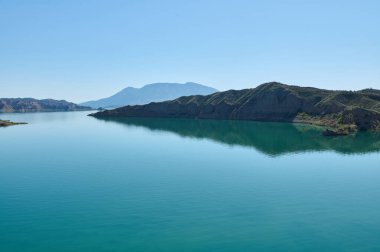 Berrak mavi gökyüzünü yansıtan dingin göl, uzakta görkemli bir dağla yeşil tepeler tarafından kucaklanıyor. Huzurlu ve manzaralı bir manzara huzuru ve doğal güzelliği çağrıştırıyor..