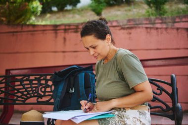 Sırt çantasıyla bankta oturan bir kadın, dikkatle kağıtlara yazı yazıyor. Serin bir ortamda çalışmanın, odaklanmanın ve çalışkan çalışmanın resmedildiği açık hava ortamı.