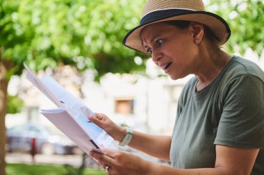 Bir kadın, açık hava ortamında abartılı bir ifade gösterirken elinde kağıtlar tutuyor. Sahne, arka planda yeşil yapraklarla duygu, odaklanma ve tepkiyi yakalar. Sıradan ve hayat dolu bir an..